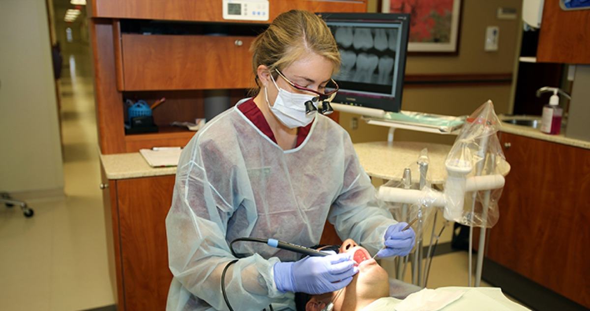 A dental hygienist in a gown, mask and loupes treating a patient